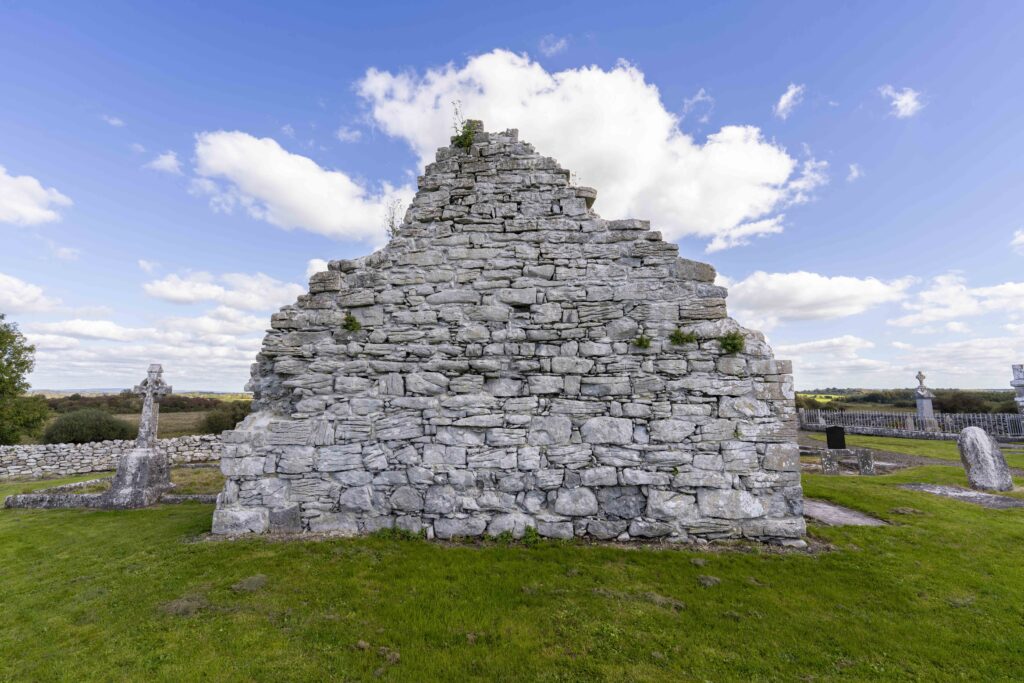 Killoo Church stone wall in a grassy area, surrounded by a cemetery with tombstones and crosses under a blue sky with scattered white clouds.