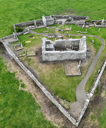 Aerial view of an ancient stone ruin enclosed by a triangular stone wall, surrounded by green grass with various stone structures and pathways within the enclosure.