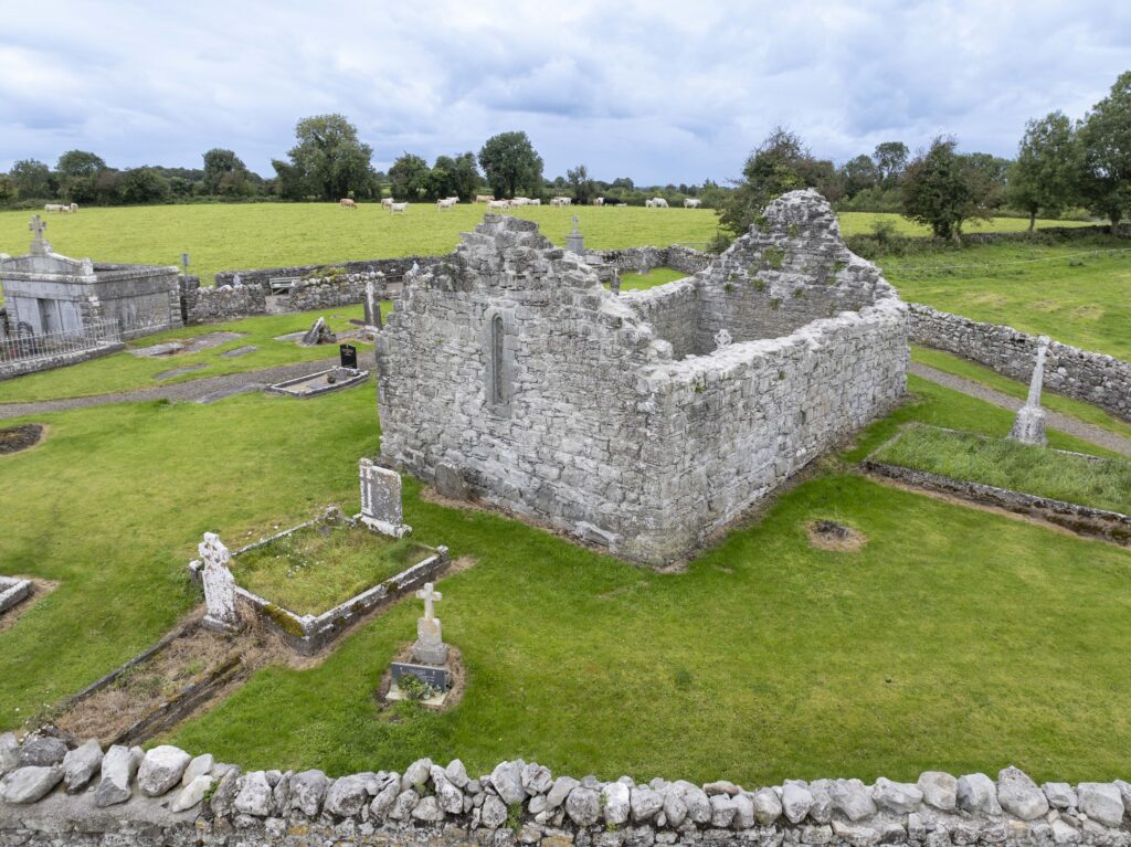 Aerial view of an old stone ruin surrounded by a graveyard with weathered tombstones and crosses, set on lush green grass and bordered by a low stone wall, with fields and trees in the background under a cloudy sky.