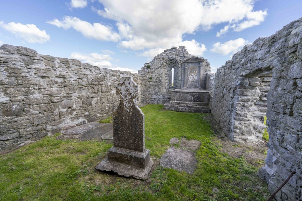 Ruins of an old stone building with weathered tombstones and grassy ground under a partly cloudy blue sky.