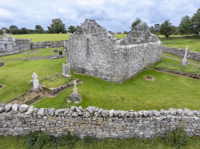 Ruined Killoo Church featuring the east window, surrounded by gravestones and lush green grass under a cloudy sky.